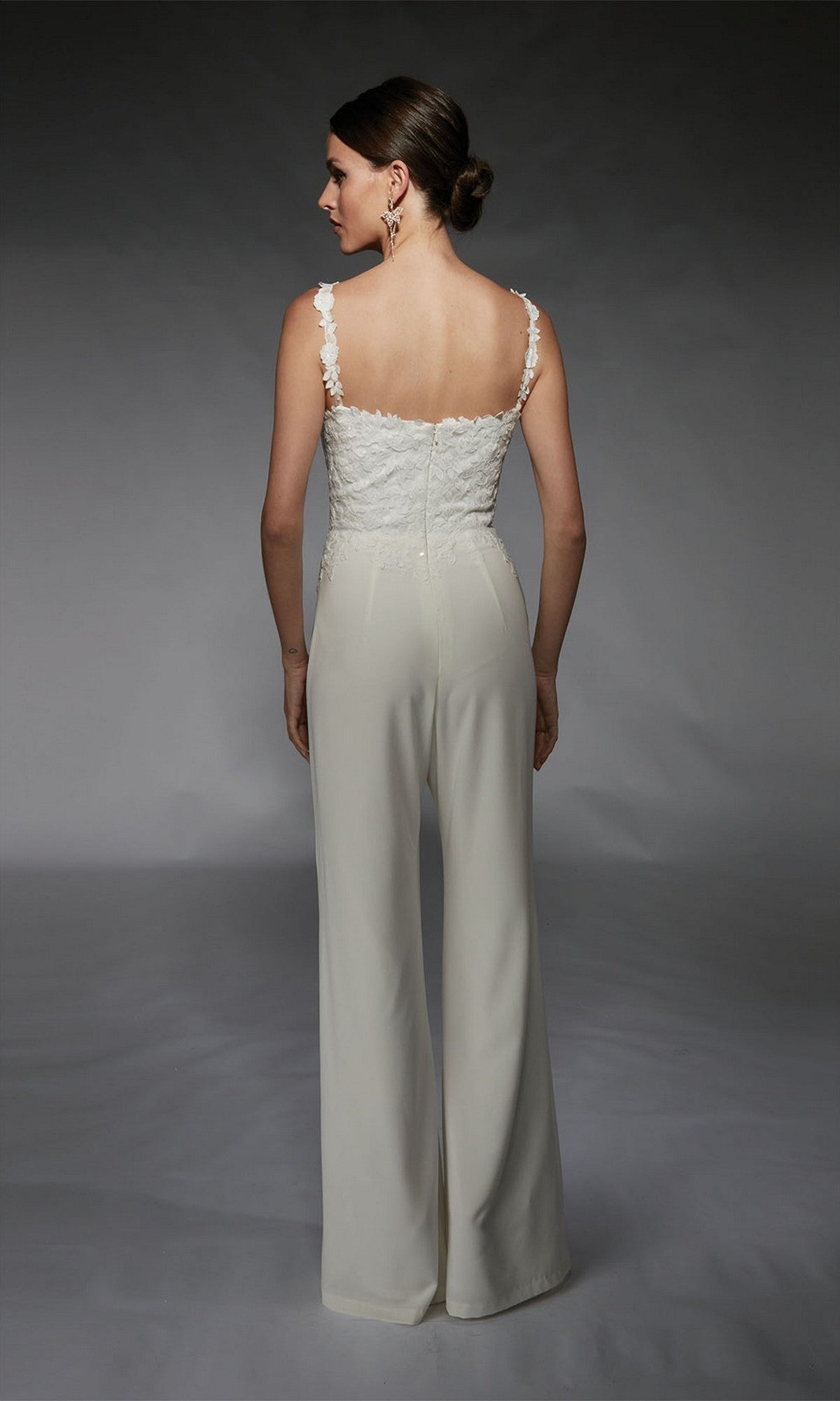 A woman faces away, wearing the Alyce 70073 Long Formal Jumpsuit—featuring a sleeveless white lace top with floral embroidery and wide-legged pants—set against a plain gray background.